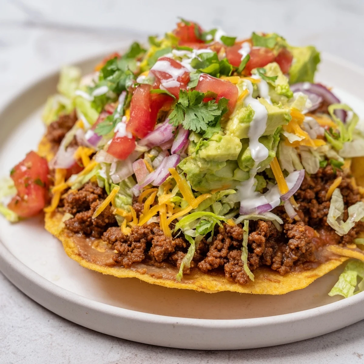 Freshly assembled Beef Tostadas with refried beans sit on a white plate, topped with diced tomatoes, shredded lettuce, and melted cheese.