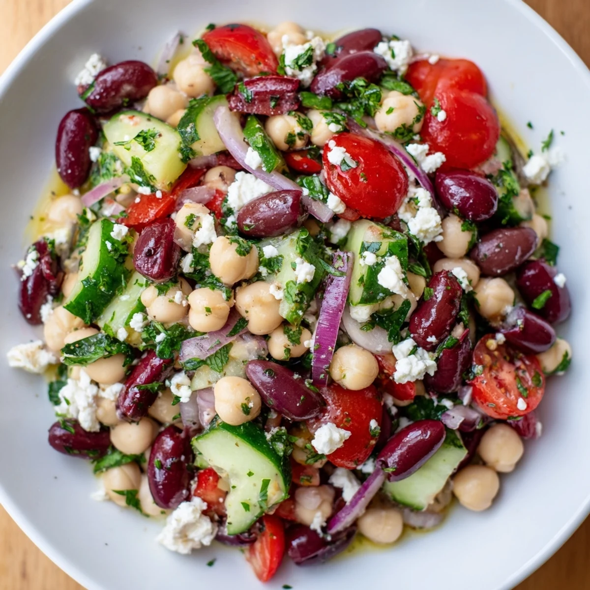 Fresh Mediterranean Dense Bean Salad in a white bowl with chickpeas, tomatoes, cucumber, olives, and crumbled feta cheese on a rustic wooden table.